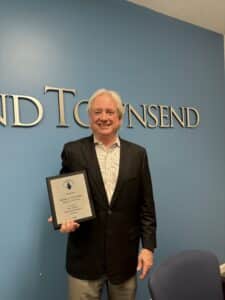 George Townsend holding "WILG top 100 workers' compensation attorney in the US 2026" award in front of law firm's sign against a blue wall.